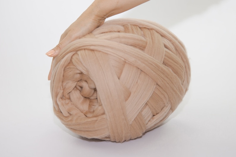 May include: A large ball of beige yarn, tightly woven and textured, is held by a hand. The yarn appears soft and thick, suitable for chunky knit projects. The neutral color makes it versatile for various crafting needs.
