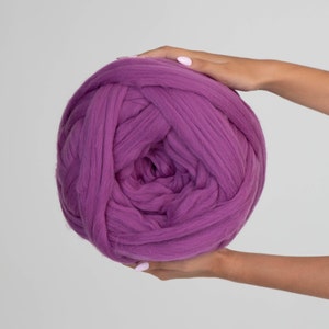 May include: A large ball of purple merino wool yarn. The yarn is thick and chunky, suitable for arm knitting or creating large, cosy projects. The ball is held by two hands against a plain white background.