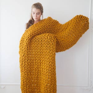 May include: A large, mustard-yellow chunky knit blanket. The blanket is made of thick yarn and has a textured appearance. The blanket is being held up, showcasing its size and warmth. The color is a rich, warm tone, perfect for home decor.