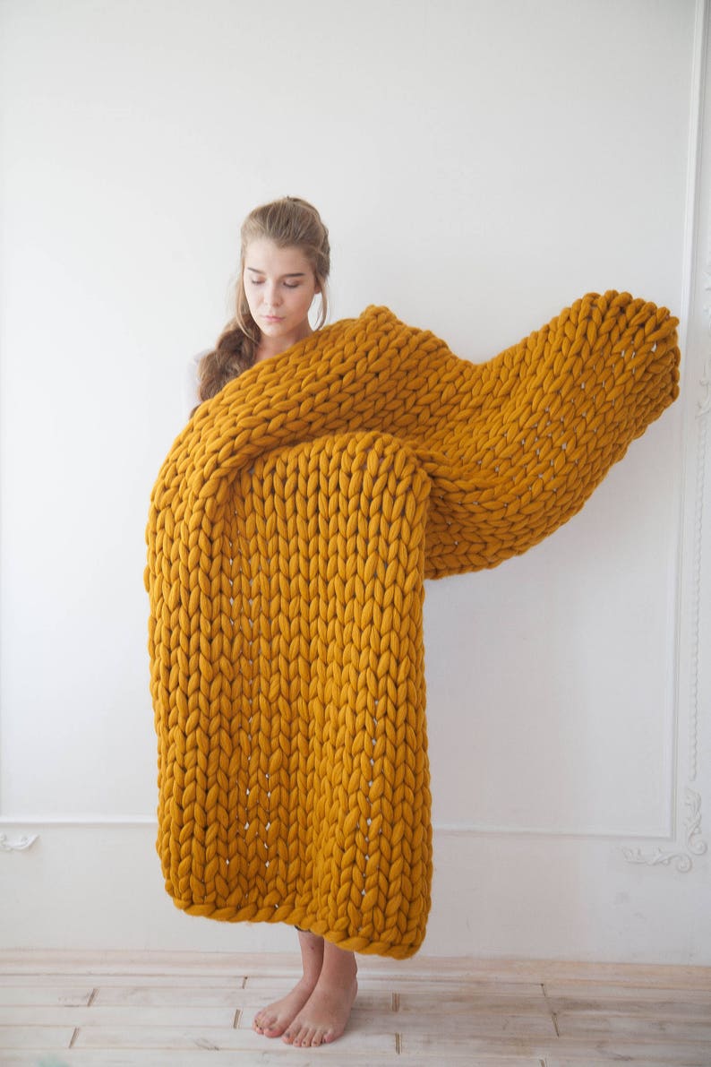 May include: A large, mustard-yellow chunky knit blanket. The blanket is made of thick yarn and has a textured appearance. The blanket is being held up, showcasing its size and warmth. The color is a rich, warm tone, perfect for home decor.