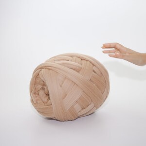 May include: A large ball of beige yarn, tightly woven with thick strands, dominates the frame. The yarn is a soft, neutral color, and the texture is clearly visible. A hand reaches towards the yarn, suggesting scale and interaction. The background is plain white.