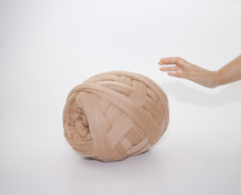 May include: A large ball of beige yarn, tightly woven with thick strands, dominates the frame. The yarn is a soft, neutral color, and the texture is clearly visible. A hand reaches towards the yarn, suggesting scale and interaction. The background is plain white.