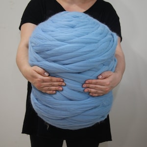 May include: A large, light blue ball of chunky knit yarn. The yarn is held by a person, showcasing its size and texture. The color is a soft, pastel blue, ideal for various crafting projects like blankets or scarves.