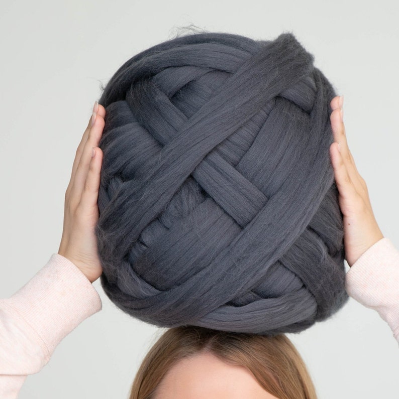 May include: A large ball of dark grey chunky knit yarn. The yarn is held by two hands, with the ball resting above the head. The yarn appears soft and thick, suitable for oversized knitting projects. The colour is a solid, neutral grey.