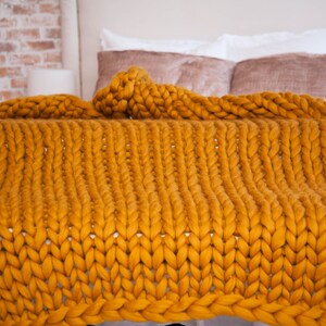 May include: A mustard yellow, chunky knit blanket draped over a bed. The blanket's thick yarn creates a textured, cozy appearance. The bed has white linens and decorative pillows, with a brick wall in the background.