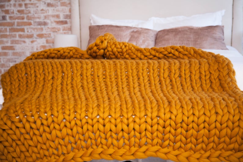 May include: A mustard yellow, chunky knit blanket draped over a bed. The blanket's thick yarn creates a textured, cozy appearance. The bed has white linens and decorative pillows, with a brick wall in the background.