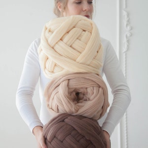 May include: Three large, chunky knit yarn balls in cream, tan, and brown hues. The yarn is thick and woven, suggesting a cozy, handmade craft project. The balls are held in front of a person, showcasing the color variations.