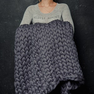 May include: A large, chunky-knit blanket in a deep grey colour. The blanket is made of thick yarn, creating a textured appearance. The words "WILDEST MOMENTS" are printed on the light grey long-sleeve shirt.