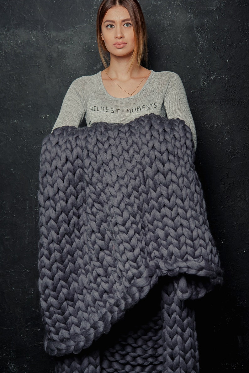 May include: A large, chunky-knit blanket in a deep grey colour. The blanket is made of thick yarn, creating a textured appearance. The words "WILDEST MOMENTS" are printed on the light grey long-sleeve shirt.