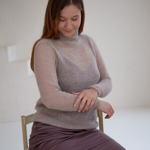 May include: A woman wearing a sheer, light brown turtleneck sweater and a mauve satin skirt. The sweater has long sleeves and a ribbed collar. She is seated on a light-colored chair in a bright room.