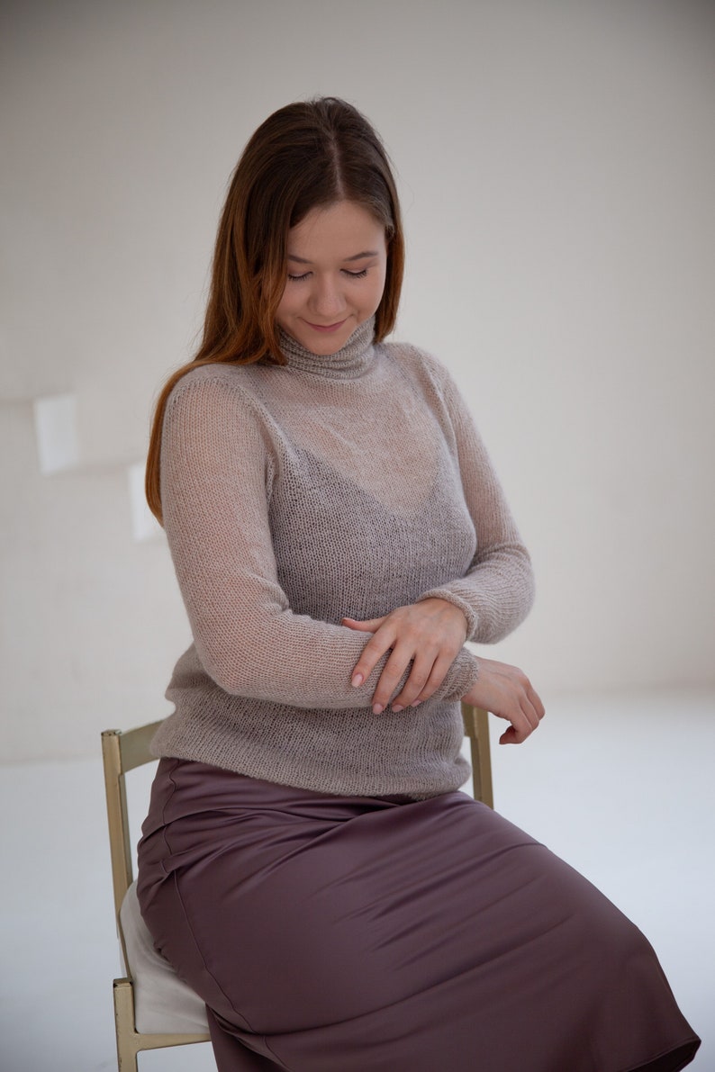 May include: A woman wearing a sheer, light brown turtleneck sweater and a mauve satin skirt. The sweater has long sleeves and a ribbed collar. She is seated on a light-colored chair in a bright room.