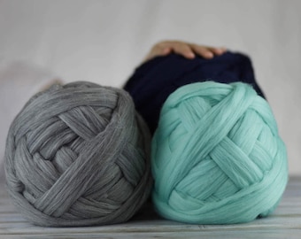 May include: Three large balls of chunky yarn in shades of gray, teal, and navy blue. The yarn is thick and appears soft, ideal for knitting or crocheting projects. The balls are arranged on a light-colored surface.