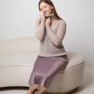 May include: A woman wearing a beige turtleneck sweater and a mauve satin midi skirt. She is seated on a cream-colored curved sofa, wearing matching pink open-toe heels. The outfit is simple and elegant.