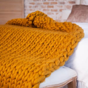 May include: A mustard yellow chunky knit blanket draped over a white bench and bed. The blanket's thick yarn creates a textured, cozy appearance. The bed has white linens and brown pillows. The background includes a brick wall and wooden panels.