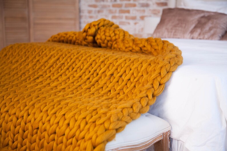 May include: A mustard yellow chunky knit blanket draped over a white bench and bed. The blanket's thick yarn creates a textured, cozy appearance. The bed has white linens and brown pillows. The background includes a brick wall and wooden panels.
