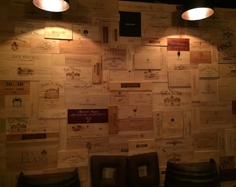 Puede incluir: Una pared cubierta de cajas de madera de vino, cada una con una etiqueta diferente. Las cajas están dispuestas en un patrón aleatorio, creando un diseño único e interesante. La pared es de color marrón claro, y las cajas son de color marrón oscuro. Hay tres sillas en primer plano, que están desenfocadas.