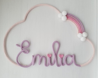 Może przedstawiać: Puchata chmurka z tęczą i imieniem "Emilia" napisanym fioletową włóczką. Tęcza ma trzy białe pompony, a imię dwa różowe serduszka.