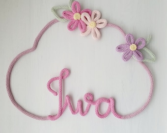 May include: A pink knitted cloud-shaped wall hanging with the name "Iwa" spelled out in pink yarn. The cloud is decorated with three crocheted flowers in pink, yellow, and purple.