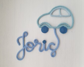 May include: A knitted wall decoration featuring a light blue car and the name "Joric" in a matching blue font. The car has two dark blue wheels. The name is in a cursive font.