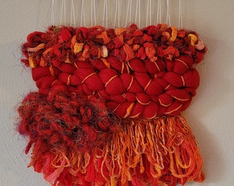 May include: A red, orange, and yellow woven wall hanging with a fringe border. The hanging is made of yarn and has a textured, braided design.