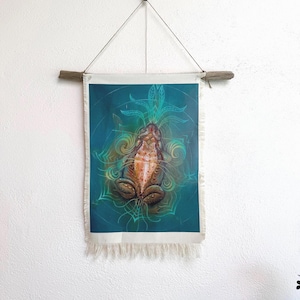 May include: A wall hanging featuring a detailed illustration of a frog. The artwork is set against a teal background with swirling patterns and botanical elements. The piece is framed by a wooden dowel and a fringe.