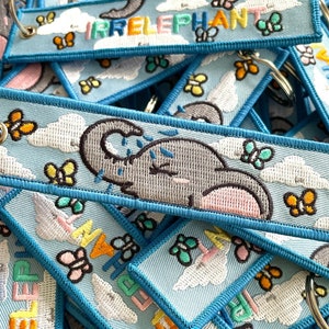Peut inclure: Un tas de porte-clés en tissu bleu et blanc avec un motif d'éléphant brodé et le texte "IRRELEPHANT". Les porte-clés présentent un fond bleu clair avec des nuages blancs et des papillons colorés.