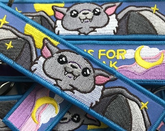 Pode incluir: Um conjunto de chaveiros azuis com morcegos bordados e uma lua crescente. Os chaveiros apresentam um esquema de cores roxo, rosa e branco e o texto "Bats for the win".