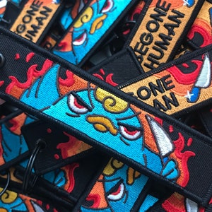 Può includere: Primo piano di diversi portachiavi colorati con un personaggio dei cartoni animati che tiene un coltello. I portachiavi sono fatti di tessuto nero con fiamme rosse, arancioni e gialle. Il testo sui portachiavi recita "BEGONE HUMAN".