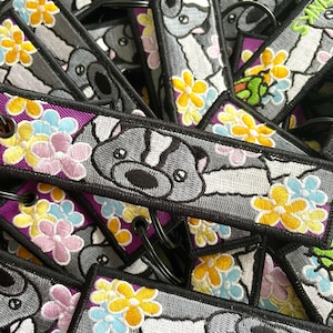 Könnte beinhalten: Nahaufnahme eines Stapels bunter Schlüsselanhänger mit einem Comic-Waschbären mit Blumen. Die Schlüsselanhänger sind aus Stoff und haben einen lila Hintergrund mit gelben, rosa und blauen Blumen.
