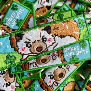Peut inclure: Porte-clés brodés avec un dessin de chien de dessin animé. Le chien a des poils bruns et blancs, des joues roses et une expression sévère. Les porte-clés ont un fond bleu avec le texte "Spikes and Spite" et des bordures vertes avec des trèfles.