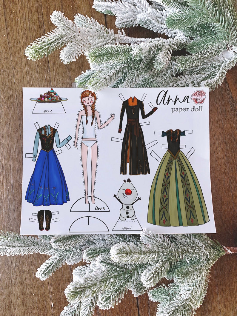 May include: A printable paper doll of Anna from the movie Frozen. The doll has a white tank top, blue skirt, and brown boots. There are several outfits to choose from, including a green dress and a black dress. The doll also comes with a stand and a snowman.