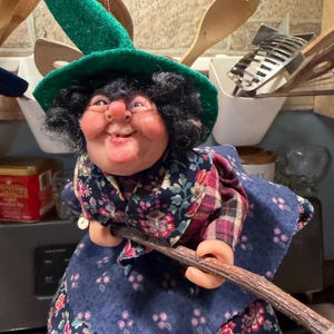 May include: A decorative doll featuring a character with black hair, a green felt hat, and a floral patterned dress. The figure has a prominent nose, small glasses, and is holding a wooden stick. The doll is dressed in a dark blue fabric garment with a red and blue plaid accent piece around the shoulders.