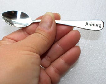 May include: A small, shiny silver spoon with the name "Ashley" engraved on the handle. The spoon has a rounded bowl and a slender handle. The spoon is held in a hand.
