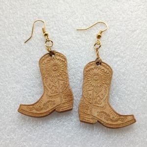 May include: A pair of wooden cowboy boot earrings with gold-colored hooks. The boots are light brown with intricate floral and scrollwork designs. Each boot features a sunflower design at the top.