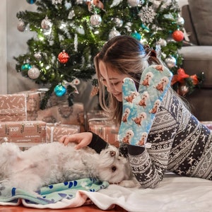 May include: A woman wearing a white and black patterned sweater with a blue oven mitt featuring a dog design. The oven mitt is on her left hand. She is petting a white dog lying on a blue and green blanket. There are wrapped gifts and a Christmas tree in the background.