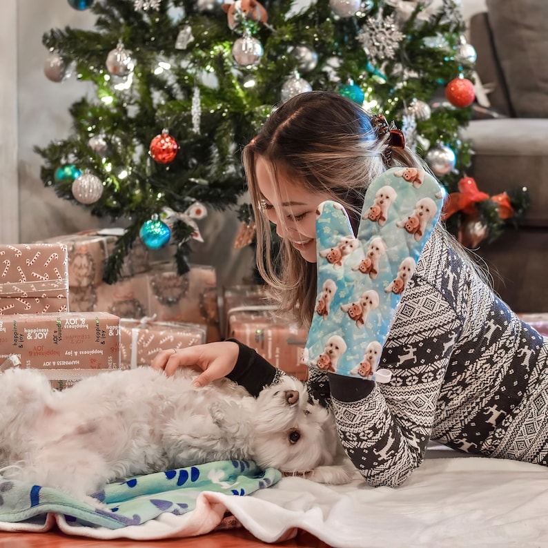 May include: A woman wearing a white and black patterned sweater with a blue oven mitt featuring a dog design. The oven mitt is on her left hand. She is petting a white dog lying on a blue and green blanket. There are wrapped gifts and a Christmas tree in the background.