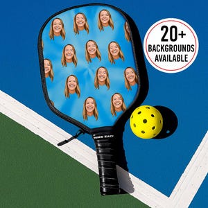 K&ouml;nnte beinhalten: Ein schwarzer Pickleball-Schl&auml;ger mit einer blauen Oberfl&auml;che, die mehrere Bilder einer l&auml;chelnden Person zeigt. Ein gelber Pickleball liegt neben dem Schl&auml;ger. Text in einem wei&szlig;en Kreis lautet "20+ BACKGROUNDS AVAILABLE".