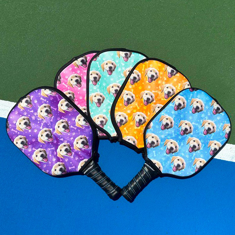K&ouml;nnte beinhalten: F&uuml;nf Pickleball-Schl&auml;ger mit schwarzen Griffen und farbenfrohen Gesichtern. Jeder Schl&auml;ger hat eine andere Hintergrundfarbe: Lila, Rosa, T&uuml;rkis, Orange und Blau. Die Schl&auml;gergesichter zeigen ein sich wiederholendes Muster aus dem Gesicht und den Knochen eines Golden Retrievers.