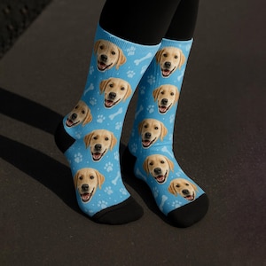 May include: Light blue socks featuring a repeating pattern of smiling golden retriever faces, white paw prints, and bone shapes. The socks have black heels and toes. The socks are worn by a person.