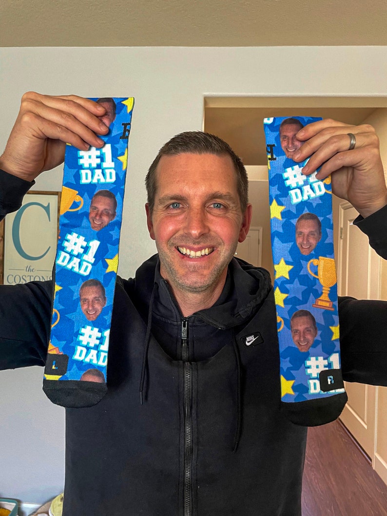 Op de afbeelding: Een paar blauwe en gele sokken met een herhalend patroon van een gezicht, een trofee en de tekst "#1 DAD".
