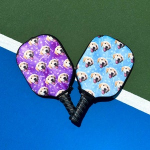 K&ouml;nnte beinhalten: Zwei Pickleball-Schl&auml;ger mit schwarzen Griffen und Designs zum Thema Hunde. Ein Schl&auml;ger hat einen lila Hintergrund mit Hundegesichtern, Knochen und Pfotenabdr&uuml;cken. Der andere hat einen blauen Hintergrund mit Hundegesichtern und Knochen.
