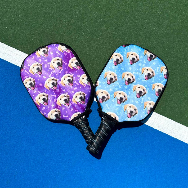 K&ouml;nnte beinhalten: Zwei Pickleball-Schl&auml;ger mit schwarzen Griffen und Designs zum Thema Hunde. Ein Schl&auml;ger hat einen lila Hintergrund mit Hundegesichtern, Knochen und Pfotenabdr&uuml;cken. Der andere hat einen blauen Hintergrund mit Hundegesichtern und Knochen.