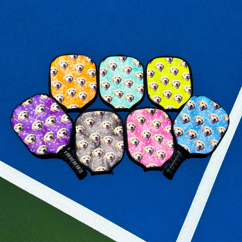K&ouml;nnte beinhalten: Eine Sammlung von Pickleball-Schl&auml;gern mit einem sich wiederholenden Muster von Golden Retriever-Gesichtern. Die Schl&auml;ger haben schwarze Griffe und sind in verschiedenen Farben erh&auml;ltlich, darunter Lila, Orange, T&uuml;rkis, Gelb, Grau, Rosa und Blau.