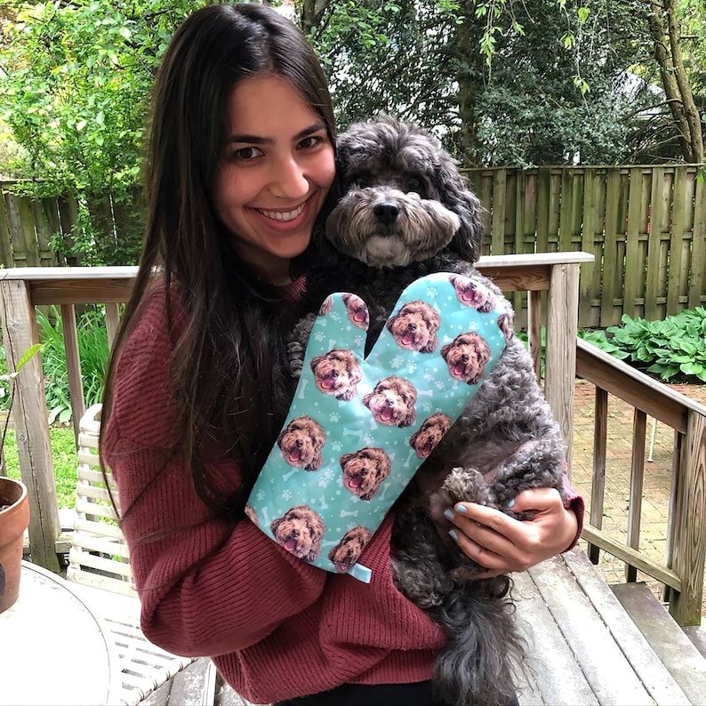 May include: A woman is holding a brown dog in her arms. She is wearing a teal oven mitt with a pattern of the dog's face on it.