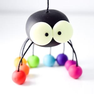 May include: A black spider with large white eyes and rainbow colored legs. The spider is made of polymer clay and is a handmade craft.
