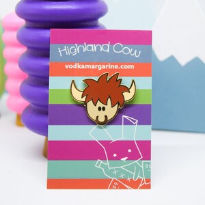 Op de afbeelding: Gouden emaille pin met een cartoon Highland Cow met een roodbruine, harige manen. De pin is op een witte kaart met de tekst "Highland Cow" en "vodkamargarine.com".