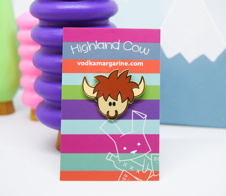 Op de afbeelding: Gouden emaille pin met een cartoon Highland Cow met een roodbruine, harige manen. De pin is op een witte kaart met de tekst "Highland Cow" en "vodkamargarine.com".