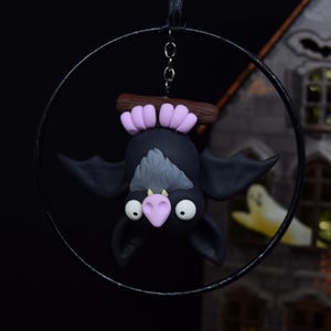 May include: A black bat ornament with a gray chest and pink accents hangs from a black metal hoop. The bat is suspended upside down, with large white eyes and a pink nose. The ornament is a Halloween decoration.