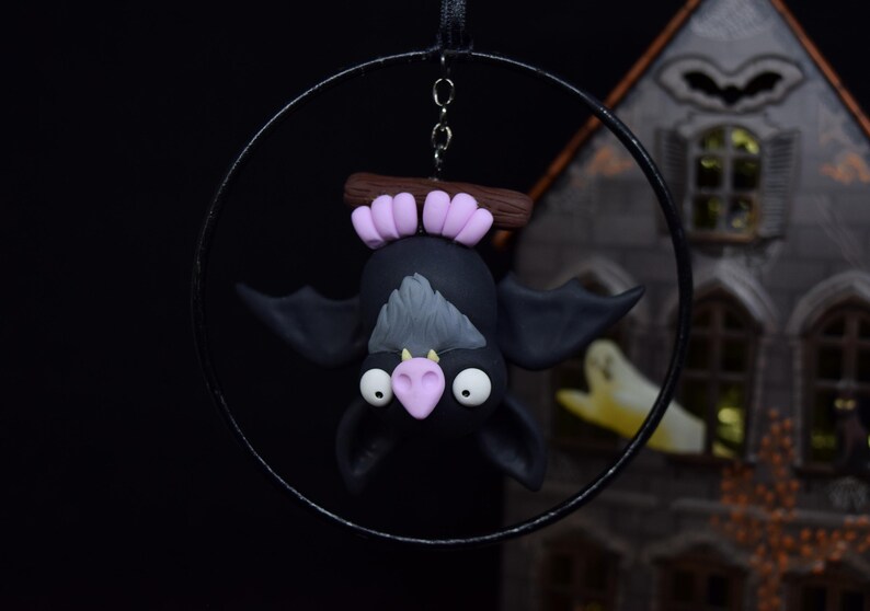 May include: A black bat ornament with a gray chest and pink accents hangs from a black metal hoop. The bat is suspended upside down, with large white eyes and a pink nose. The ornament is a Halloween decoration.