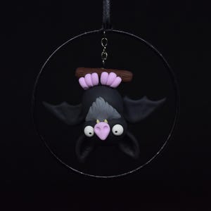 May include: A whimsical, handmade bat ornament. The bat is black with gray accents, large white eyes, and a pink nose and ears. It hangs upside down from a brown branch with pink feet, suspended within a metal ring. The background is solid black.